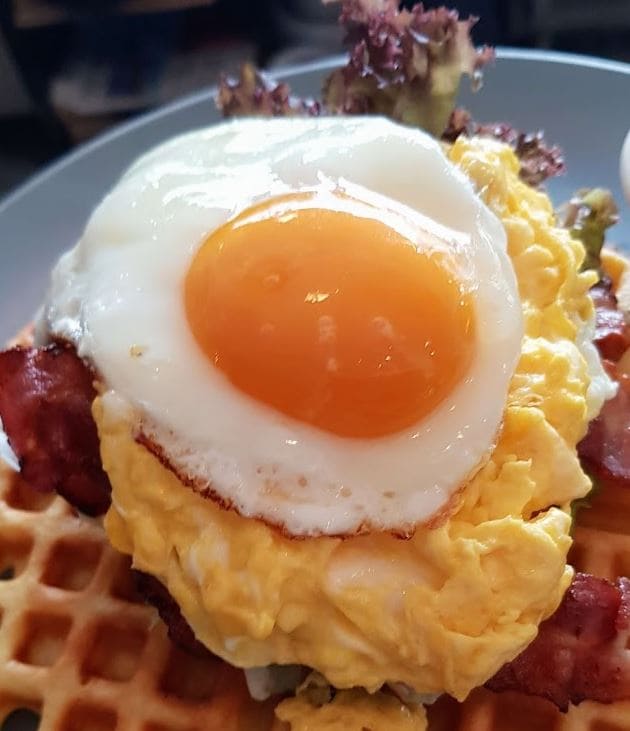 sunny side up egg on scrambled eggs and bacon with salad on top of waffle piece